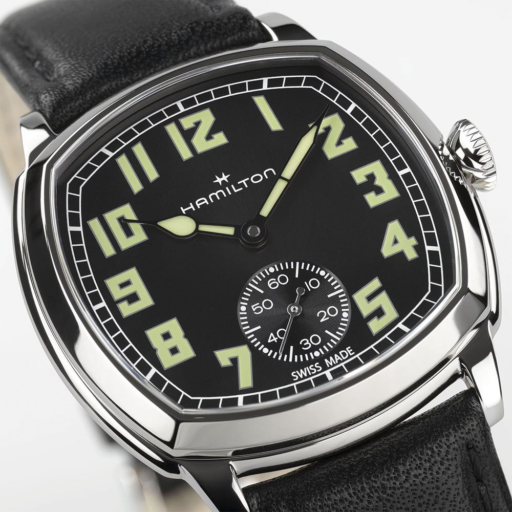 Angled view of American Classic Cushion Mechanical 34mm watch with black dial luminous numerals and polished steel case on a black leather strap.