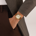 Gold-tone cushion wristwatch with beige leather strap worn on a wrist beneath a gray suit jacket.