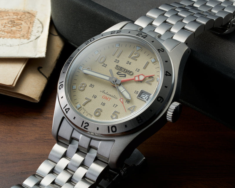 A Seiko stainless steel wristwatch with a silver link bracelet, featuring a beige dial, black bezel, and a red seconds hand, displayed on a dark surface next to a watch box.